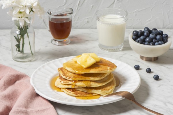 Buttermilk pancakes with syrup and butter, accompanied by milk and berries, set against a backdrop of white flowers on a marble countertop